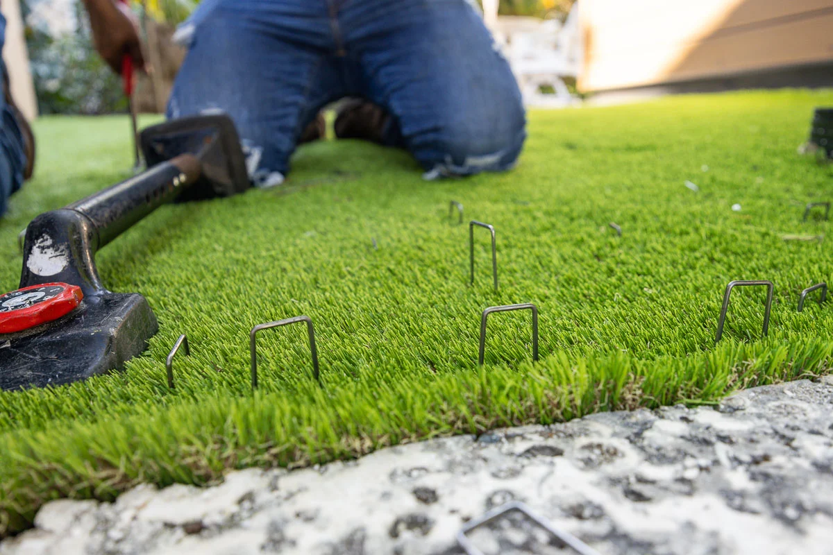 Worker kneeling to secure artificial turf with landscape staples during a front yard landscaping installation in the San Fernando Valley