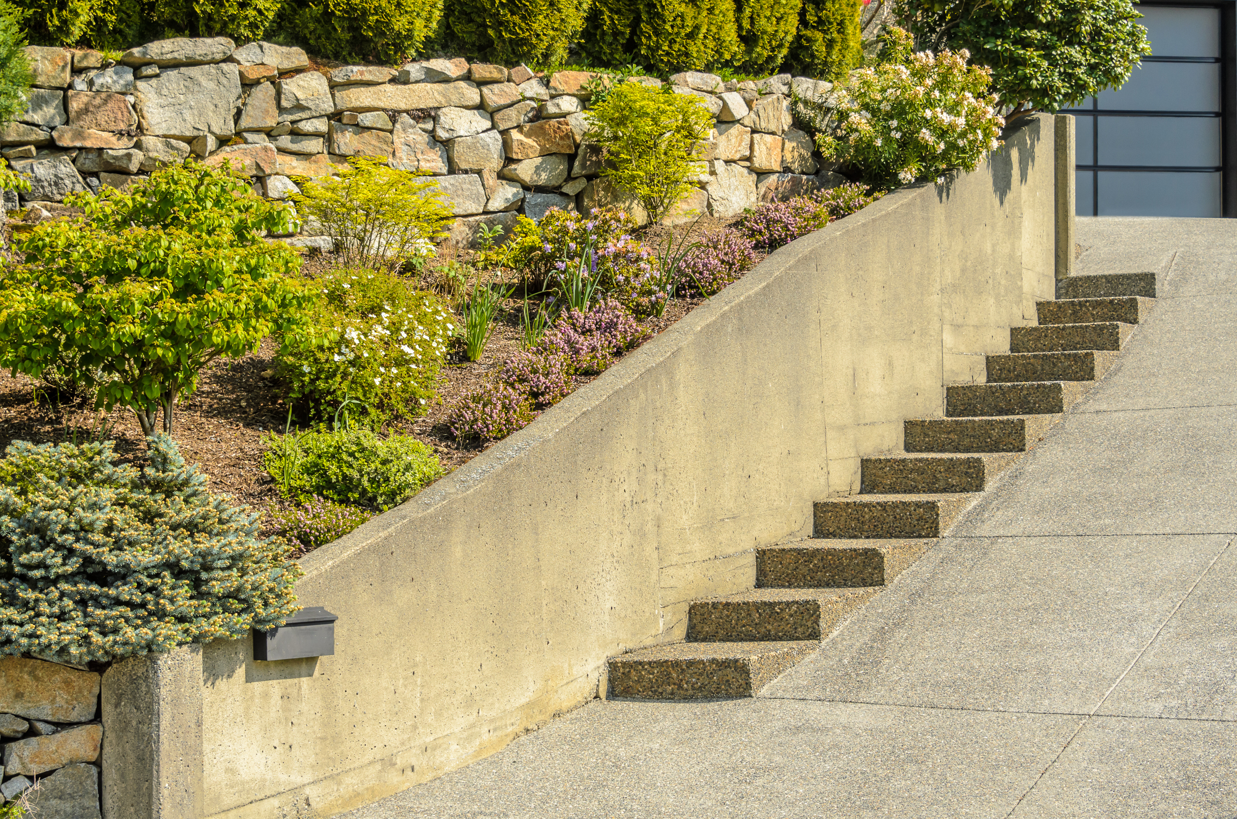 Hardscape design with a stone retaining wall, planted slope, and concrete steps leading up to a driveway in the San Fernando Valley